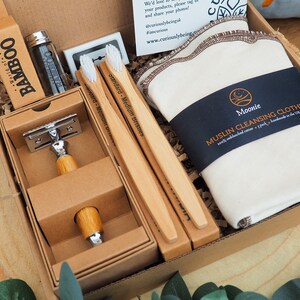 May include: A brown cardboard box filled with a variety of natural bath and body products. The box contains a bamboo toothbrush, a safety razor, a muslin cleansing cloth, and a bottle of floss. The box is labeled "World of Bamboo".