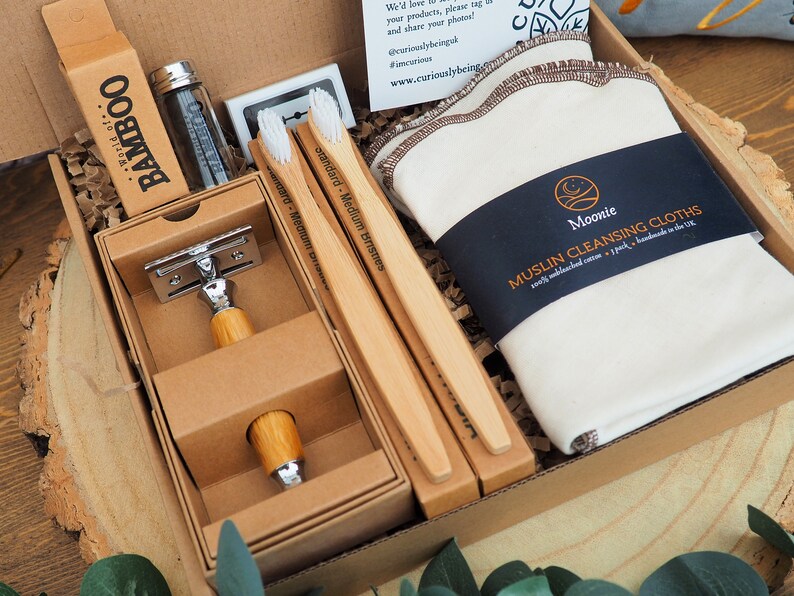 May include: A brown cardboard box filled with a variety of natural bath and body products. The box contains a bamboo toothbrush, a safety razor, a muslin cleansing cloth, and a bottle of floss. The box is labeled "World of Bamboo".