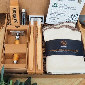 May include: A brown cardboard box filled with a zero waste gift set. The set includes a bamboo razor, two bamboo toothbrushes, a glass jar with a metal lid, and a white muslin cleansing cloth with a black label that says "Moonie Muslin Cleansing Cloths".