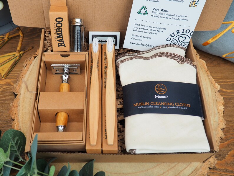 May include: A brown cardboard box filled with a zero waste gift set. The set includes a bamboo razor, two bamboo toothbrushes, a glass jar with a metal lid, and a white muslin cleansing cloth with a black label that says "Moonie Muslin Cleansing Cloths".