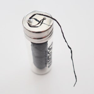 May include: A small glass jar with a silver lid containing black dental floss. The floss is pulled out of the jar and is hanging down.