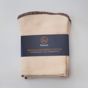 May include: A stack of 7 natural colored muslin cleansing cloths with brown stitching. The cloths are packaged with a black label that reads "Moonie Muslin Cleansing Cloths 100% unbleached cotton - 7 pack - handmade in the UK"