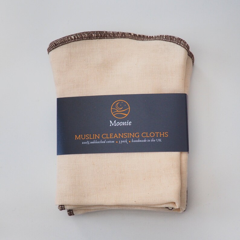 May include: A stack of 7 natural colored muslin cleansing cloths with brown stitching. The cloths are packaged with a black label that reads "Moonie Muslin Cleansing Cloths 100% unbleached cotton - 7 pack - handmade in the UK"