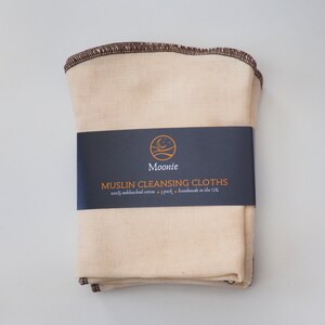 K&ouml;nnte beinhalten: Ein Stapel von 7 naturfarbenen Musselin-Reinigungst&uuml;chern mit braunen N&auml;hten. Die T&uuml;cher sind mit einem schwarzen Etikett verpackt, auf dem "Moonie Muslin Cleansing Cloths 100% unbleached cotton - 7 pack - handmade in the UK" steht.