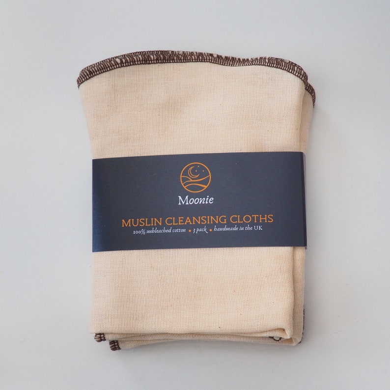 K&ouml;nnte beinhalten: Ein Stapel von 7 naturfarbenen Musselin-Reinigungst&uuml;chern mit braunen N&auml;hten. Die T&uuml;cher sind mit einem schwarzen Etikett verpackt, auf dem "Moonie Muslin Cleansing Cloths 100% unbleached cotton - 7 pack - handmade in the UK" steht.