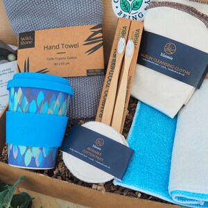 K&ouml;nnte beinhalten: Eine Geschenkbox gef&uuml;llt mit umweltfreundlichen Produkten, darunter eine Bambuszahnb&uuml;rste, wiederverwendbare Wattepads, ein Handtuch, eine wiederverwendbare Kaffeetasse und Mull-Reinigungst&uuml;cher. Die Box ist mit einem gr&uuml;nen Blatt und den Worten "Bring Your Being" verziert.