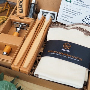 May include: A brown cardboard box filled with eco-friendly bathroom essentials. The box contains a bamboo razor, three bamboo toothbrushes, and a pack of white muslin cleansing cloths. The box is labeled "Moonie Muslin Cleansing Cloths" and "Curiously Being".