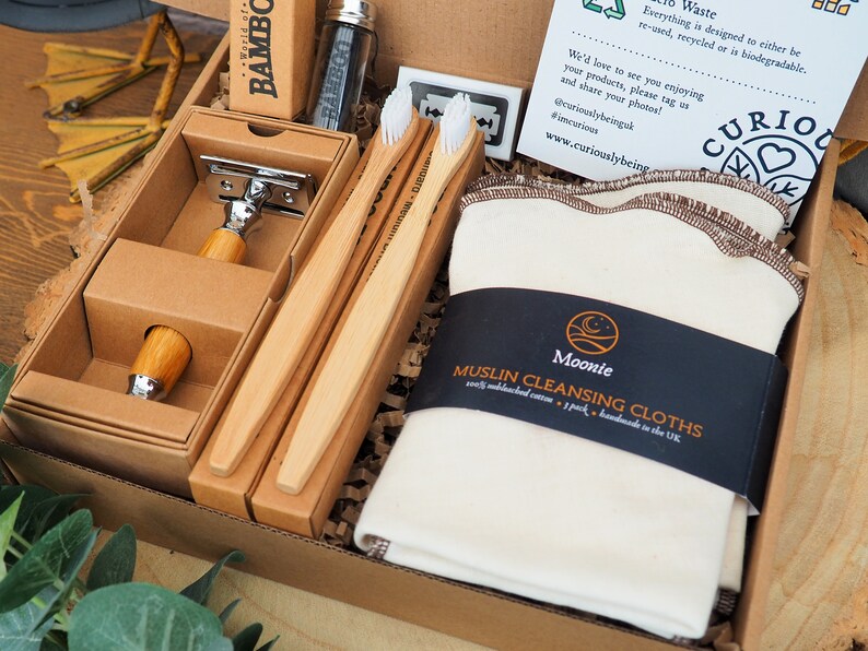 May include: A brown cardboard box filled with eco-friendly bathroom essentials. The box contains a bamboo razor, three bamboo toothbrushes, and a pack of white muslin cleansing cloths. The box is labeled "Moonie Muslin Cleansing Cloths" and "Curiously Being".
