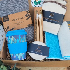 K&ouml;nnte beinhalten: Eine braune Pappbox gef&uuml;llt mit umweltfreundlichen Bad- und K&uuml;chenutensilien. Die Box enth&auml;lt eine blaue wiederverwendbare Kaffeetasse, zwei Bambuszahnb&uuml;rsten, ein graues Handtuch, ein wei&szlig;es Musselin-Reinigungstuch, ein blaues Reinigungstuch und eine Packung wiederverwendbarer Wattepads.