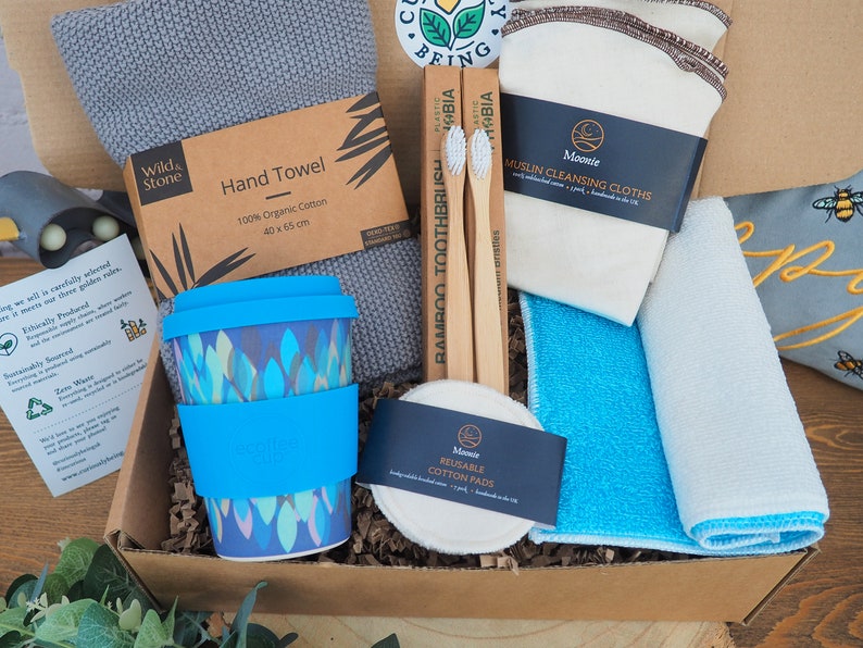 K&ouml;nnte beinhalten: Eine braune Pappbox gef&uuml;llt mit umweltfreundlichen Bad- und K&uuml;chenutensilien. Die Box enth&auml;lt eine blaue wiederverwendbare Kaffeetasse, zwei Bambuszahnb&uuml;rsten, ein graues Handtuch, ein wei&szlig;es Musselin-Reinigungstuch, ein blaues Reinigungstuch und eine Packung wiederverwendbarer Wattepads.