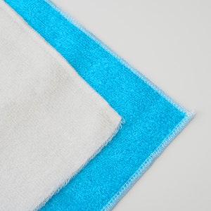 K&ouml;nnte beinhalten: Zwei Mikrofaser-T&uuml;cher, eines wei&szlig; und eines blau, sind zusammengelegt. Die T&uuml;cher haben einen gen&auml;hten Rand.
