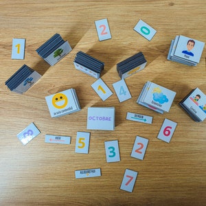 May include: A set of colorful magnetic cards for teaching children about days of the week, months, and numbers. The cards feature images of a smiling face, a cloud with a sun, a tree, a person, and the words "hier", "demain", "aujourd'hui", "octobre", and "eclecies".