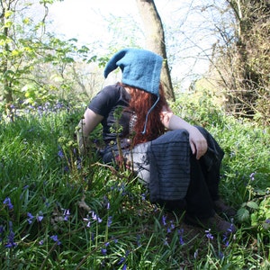 Puede incluir: Una persona con un gorro de punto azul se sienta en un campo de campanillas. El gorro tiene una punta larga y puntiaguda y está tirado sobre la cabeza de la persona. La persona lleva una camisa negra y pantalones oscuros. Las campanillas están en flor y son de un color azul vibrante.