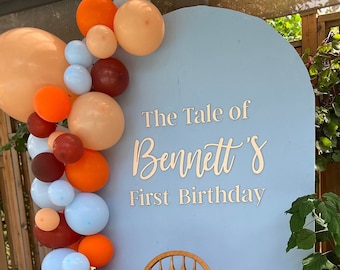 May include: A blue backdrop with the text "The Tale of Bennett's First Birthday" in white. A balloon arch made of orange, brown, and blue balloons is on the left side of the backdrop. A wooden high chair with a brown and white party hat is in front of the backdrop.