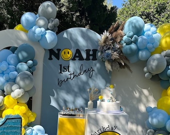 May include: A blue and yellow balloon arch with a white and blue backdrop. The backdrop has the text "Noah 1st birthday" and a smiley face. There is a blue and yellow cake on a white table with a yellow and white cake stand. There is a large blue number one with a smiley face and the text "Noah" on it.