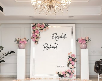 May include: A white backdrop with a silver frame and pink flowers on either side. The backdrop has the text "Elizabeth + Richard" and the date "24.10.2023" written in black.
