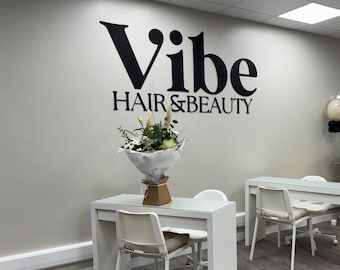 May include: A white reception desk with a round mirror featuring the word "Vibe" in black lettering. Two white tables with chairs are in the foreground. The wall behind the desk has a black and white sign that reads "Vibe Hair & Beauty".