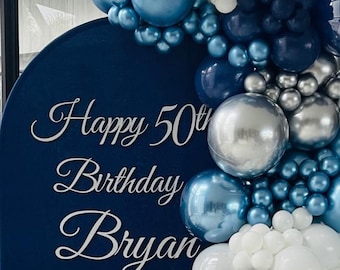 May include: A blue backdrop with the words "Happy 50th Birthday Bryan" in silver lettering. The backdrop is decorated with a garland of blue, silver, and white balloons.