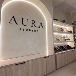 以下が含まれることがあります： 「AURA STUDIOS」の文字が黒で書かれたスタジオの内観。壁は明るいベージュで、曲線を描く照明付きのアーチがあります。木製のキャビネットと棚には、マニキュアボトルなど、さまざまなアイテムが展示されています。
