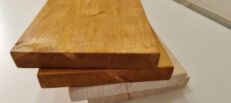 May include: Three wooden boards stacked on top of each other. The top board is stained a dark orange color, while the bottom two boards are a natural wood color.