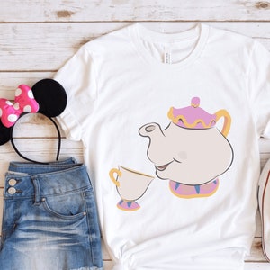 Puede incluir: Camiseta blanca con una imagen de dibujos animados de la Sra. Tetera, la tetera de La Bella y la Bestia, sirviendo té en una taza.