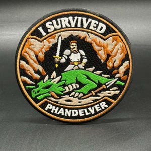 May include: Round patch with the text "I SURVIVED PHANDELVER". The patch features a knight with a sword standing over a green dragon. The background includes brown rock formations.
