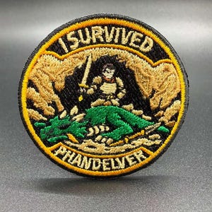 May include: A circular embroidered patch featuring a black background with a gold border. The text I SURVIVED PHANDELVER is stitched in gold. The center depicts a knight in armor holding a sword standing over a green dragon in a cave.