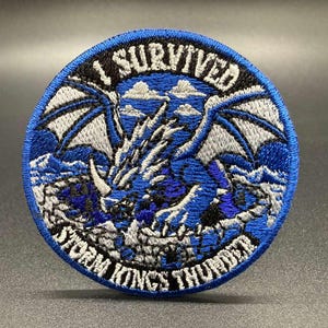 May include: A circular embroidered patch featuring a blue and white dragon with wings spread. The text I SURVIVED is arched at the top, and STORM KINGS THUNDER is arched at the bottom in white lettering against a black background border.