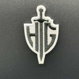 May include: Embroidered patch featuring a shield design with a sword and the letters "HIG" in black and white. The patch has a white border and is likely for clothing or accessories.