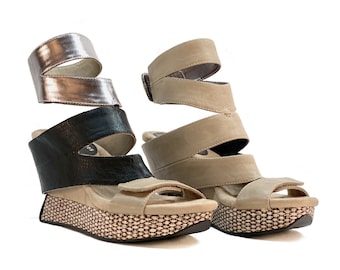 May include: Two pairs of women's wedge sandals. One pair is black and tan, the other is silver and tan. The sandals have a woven platform sole and multiple straps that wrap around the ankle and foot.
