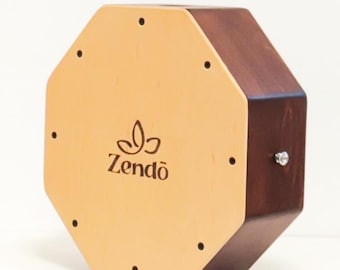 May include: An octagonal wooden percussion instrument with a light tan front panel and dark brown sides. The front panel features the word "Zendō" in a stylized font. Small black dots are evenly spaced around the front panel.