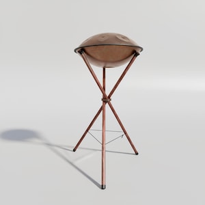 Könnte beinhalten: Ein braunes Handpan aus Stahl mit dunklem Rand, das auf einem kupferfarbenen Dreibeinständer steht. Das Handpan hat mehrere kreisförmige Vertiefungen auf seiner Oberfläche. Der Ständer hat drei Beine und eine kreisförmige Strebe.