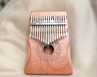 May include: A wooden kalimba with metal tines. The instrument has a reddish-brown finish and a circular design etched into the wood. The metal tines are arranged in two rows. The kalimba is a musical instrument.