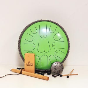 May include: A green steel tongue drum with a gray border, black numbers, and cut-out shapes. Accessories include a wooden mallet, a small metal drum, a wooden box with a leaf design, and two drumsticks. The drum is a musical instrument.