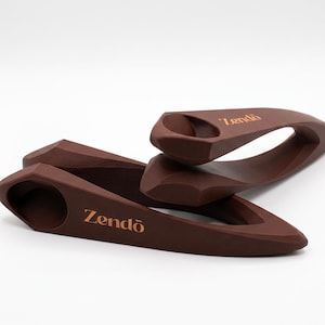 May include: Two dark brown, wedge-shaped door stops with a smooth, matte finish. Each object features a circular indentation at the wider end and the word "Zendō" printed in a light copper-colored font on the side of the tapered body.