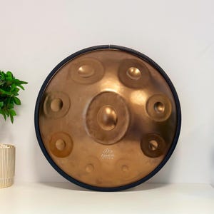 May include: A circular copper-toned handpan musical instrument with a dark blue rim. The surface features multiple circular indentations arranged around a central dome. The logo Zendo Sounds is printed in white near the bottom center of the instrument.