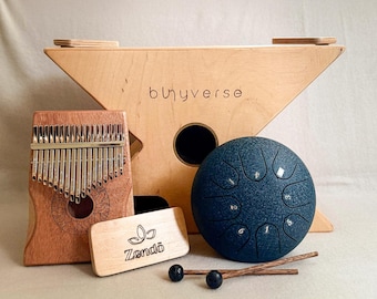 May include: A collection of musical instruments including a wooden kalimba, a blue steel tongue drum, a wooden stand with the text "bluyverse", a wooden sound block with the text "Zendō", and two wooden drumsticks with black ball ends.