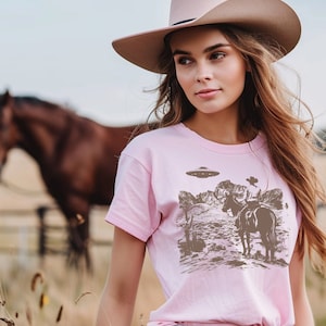 Könnte beinhalten: Eine Frau trägt ein pinkfarbenes T-Shirt mit einem Aufdruck eines Cowboys, der ein Pferd reitet, mit einem UFO im Hintergrund. Sie trägt einen braunen Cowboyhut und eine blaue Jeans.