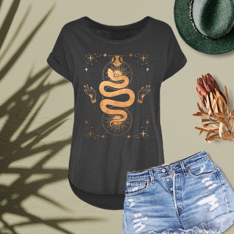 Peut inclure: Un t-shirt gris fonc&eacute; avec un motif de serpent dor&eacute;. Le serpent est enroul&eacute; autour d'un symbole de soleil et de lune. Le motif est entour&eacute; d'&eacute;toiles.