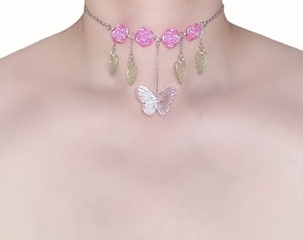 Peut inclure: Un collier ras du cou argenté orné de roses roses et de breloques en forme de feuilles vertes pendantes. Un pendentif papillon transparent est suspendu au centre. Le collier est conçu pour être porté autour du cou.
