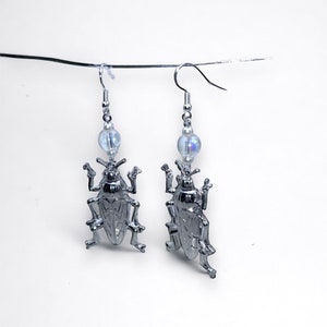 Può includere: Un paio di orecchini d'argento con un design a scarabeo e perline di vetro iridescenti.