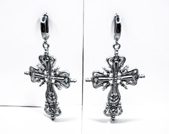 May include: A pair of silver cross earrings with intricate detailing. The earrings are dangle style and have a hoop closure.