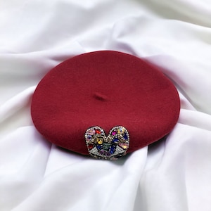May include: A burgundy beret with a colorful jeweled butterfly embellishment. The hat is round and made of a soft material. The butterfly is made of various colored gems and beads.