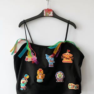 Könnte beinhalten: Ein schwarzes Camisole-Top mit dünnen Spaghettiträgern und befestigten, bunten, transparenten Bändern. Die Vorderseite ist mit gestickten Aufnähern eines Clowns, eines Tigers, eines Riesenrads, eines Elefanten, eines Karussells und dem Wort Circus verziert.