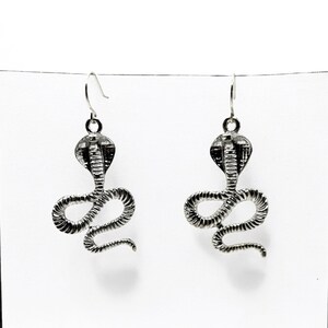 May include: A pair of silver snake earrings with a detailed design. The earrings are hanging from a silver hook.