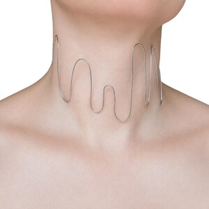 May include: A silver-toned wire choker necklace featuring a continuous, fluid, wave-like design that wraps around the neck. The metal wire forms a series of irregular, rounded loops of varying heights, creating an open, sculptural, and linear aesthetic.