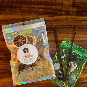 Può includere: Un sacchetto di plastica trasparente pieno di erbe secche, spezie e scorze di agrumi. Il sacchetto ha un'etichetta che recita "Abundance Simmer Pot Kit". Ci sono anche tre bastoncini di incenso e una piccola borsa verde con cristalli e un motivo a stelle.