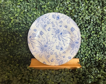May include: A white ceramic plate with a blue floral pattern. The plate is displayed on a small wooden stand against a backdrop of green foliage. The floral design includes various flowers and foliage.