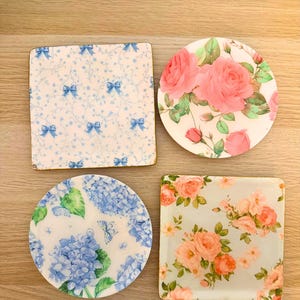 Può includere: Quattro sottobicchieri con forme e motivi vari. Un sottobicchiere quadrato presenta una stampa con fiocchi blu e viticci su sfondo bianco. Un sottobicchiere rotondo mostra rose rosa e foglie verdi. Un sottobicchiere rotondo mostra ortensie blu.