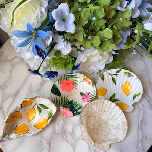 Pode incluir: Uma coleção de bases decorativas de conchas. As bases apresentam vários designs, incluindo limões, flores tropicais e um padrão floral dourado. As conchas estão dispostas sobre uma superfície de mármore, com um buquê de flores ao fundo.